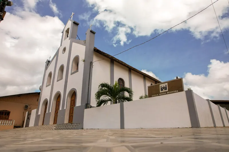 Igreja Nossa Senhora da Assunção seen diagonally, with a white wall, name plaque and a palm tree, under a blue sky with clouds.