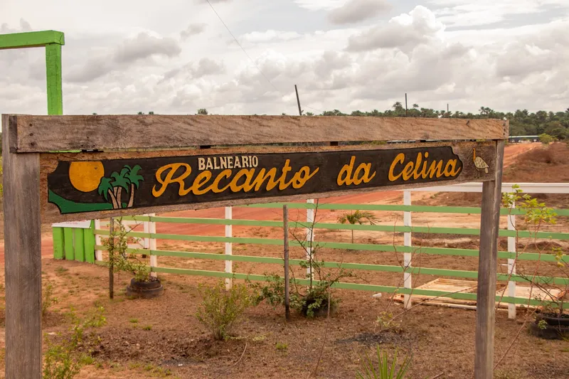 Letrero de madera en la entrada del Balneário Recanto da Celina, con cerca verde y blanca y suelo de tierra