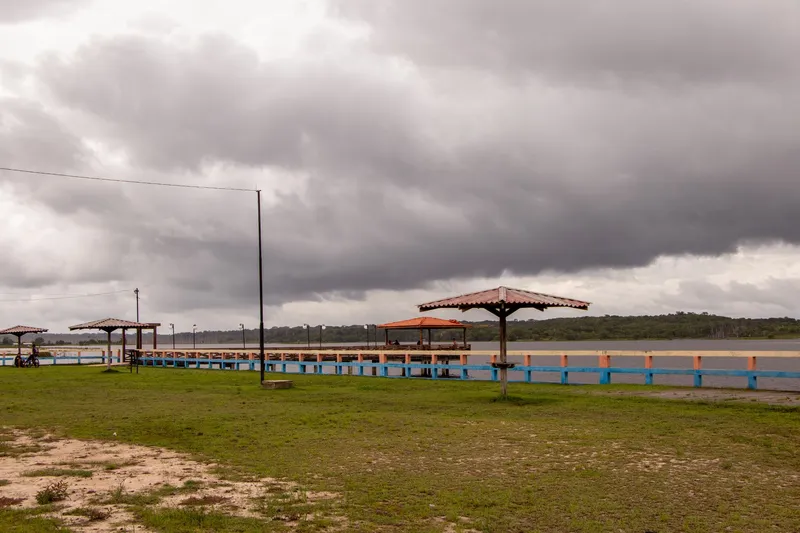 Quiosques de telhado vermelho no gramado da Orla de Porto Grande, mureta colorida e rio ao fundo sob nuvens escuras.