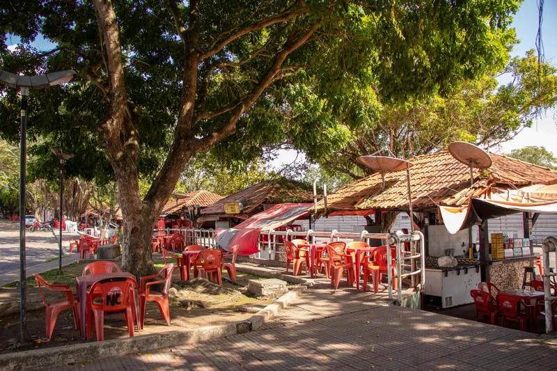 Quiosques com telhado de cerâmica e mesas vermelhas espalhadas sob árvores na Praça Isaac Zagury, com luz de sol da manhã.