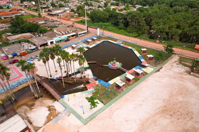 Vista aérea del balneario de Porto Grande: lago con isla central y pasarelas, quioscos coloridos, palmeras y zona de arena junto a la ciudad