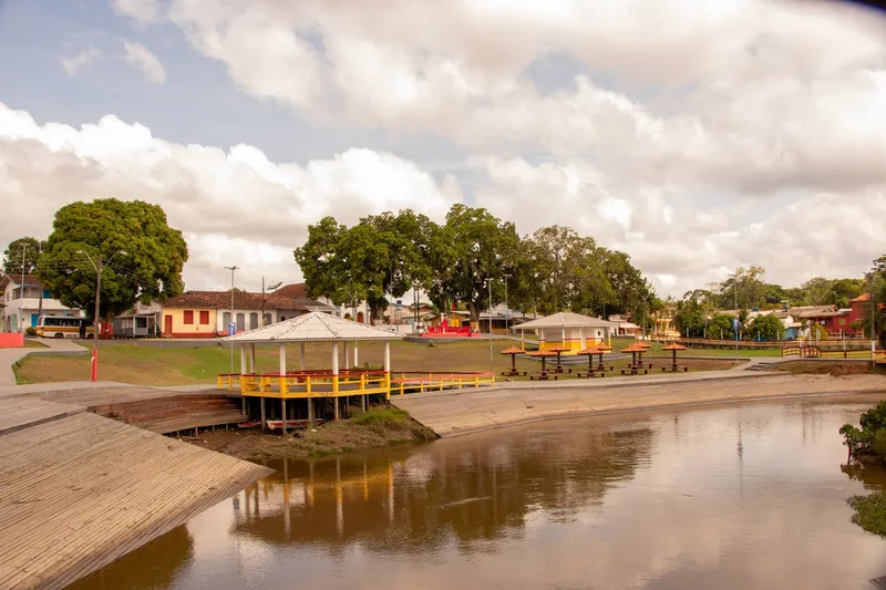 Orla de Mazagão Velho com coreto amarelo à beira do rio, mesas cobertas, gramado e casas ao fundo refletidas na água.