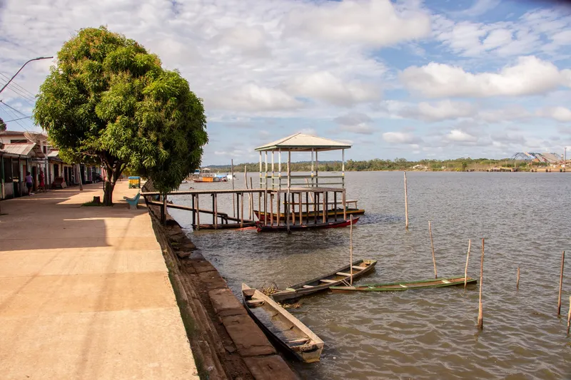 Promenade of the Vitória do Jari waterfront with a leafy tree, a covered jetty and wooden canoes on the river.
