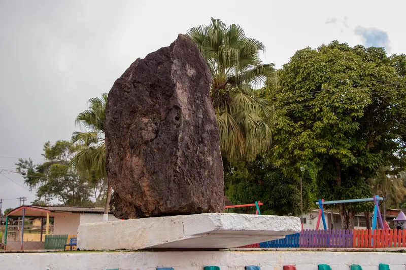 Grande rocha escura exposta sobre pedestal branco na Vila Serra do Navio, com palmeiras, árvores e parquinho colorido ao fundo.