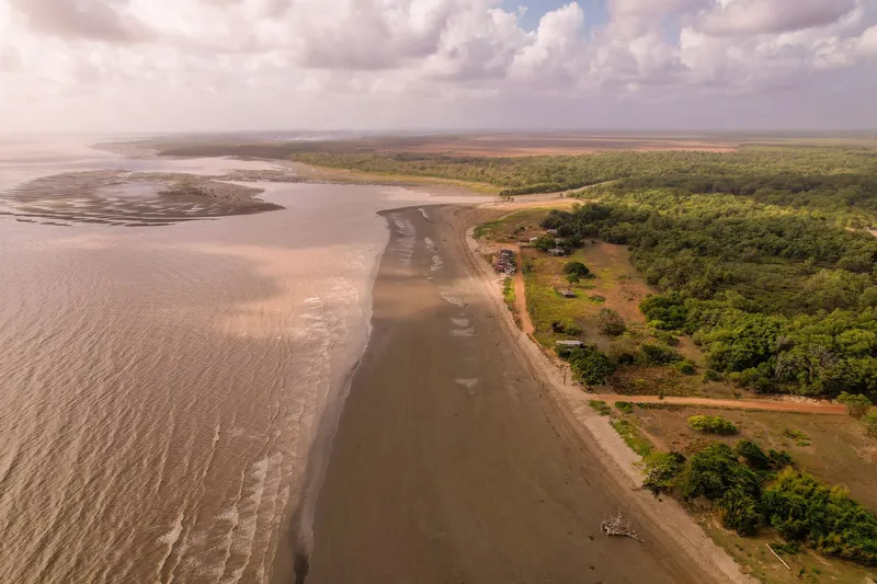 Vista aérea da Praia do Goiabal: longa faixa de areia escura entre o mar barrento e a vegetação, com casas e estrada de terra junto à orla