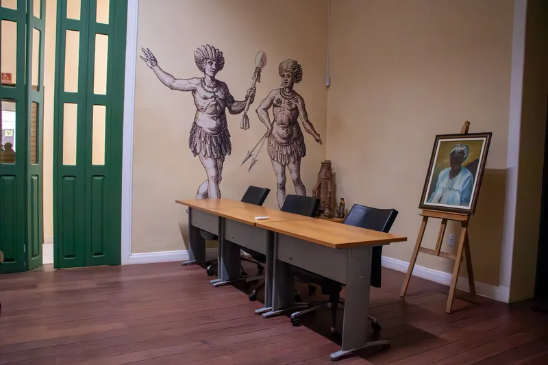 Sala del Museu Joaquim Caetano da Silva con mesas y sillas, un grabado de dos indígenas pintado en la pared y un retrato sobre un caballete.