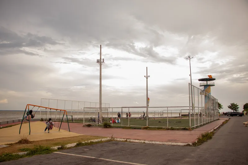 Balanços, quadra de futebol cercada e um mirante colorido na Orla do Bairro Cidade Nova, sob céu carregado, com o rio à esquerda.