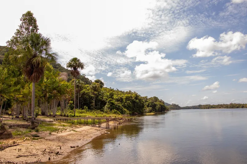 Sandy bank of Balneário Recanto do Cupu with palm trees and a small wooden pier, beside the wide river under a blue sky with clouds.