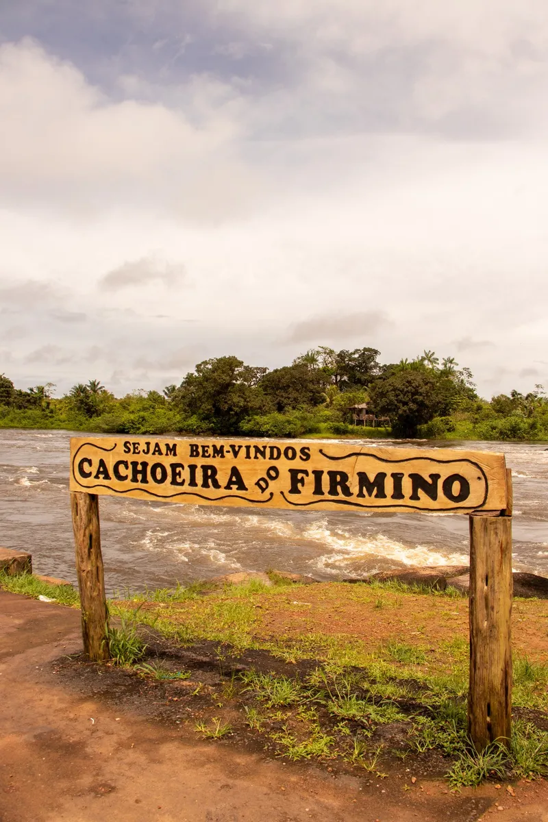 Panneau en bois « Sejam bem-vindos Cachoeira do Firmino » devant la rivière agitée, avec rapides et forêt sur l'autre rive
