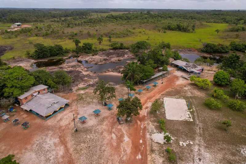 Aerial view of Balneário Asa Aberta: kiosks, hall and a dirt road beside the river with rock slabs, surrounded by grassland and forest