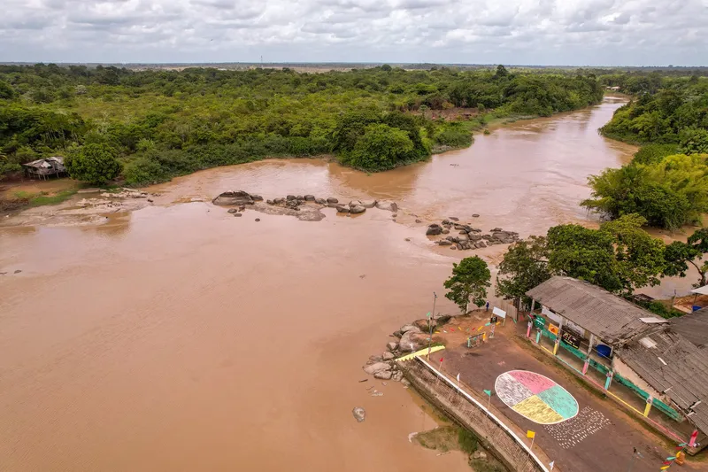 Vue aérienne de la Cachoeira do Firmino : rivière boueuse aux rochers affleurants, rives boisées et espace de loisirs coloré avec belvédère au bord de l'eau