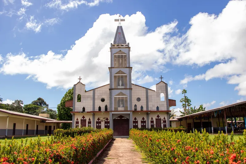 Facade of the Ilha de Santana church, with a central tower and cross, seen at the end of a path between flower beds.