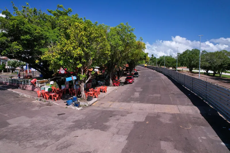 Rua em curva ao lado da Praça Isaac Zagury, com quiosques e mesas de plástico vermelhas sob grandes árvores, em dia de céu azul.
