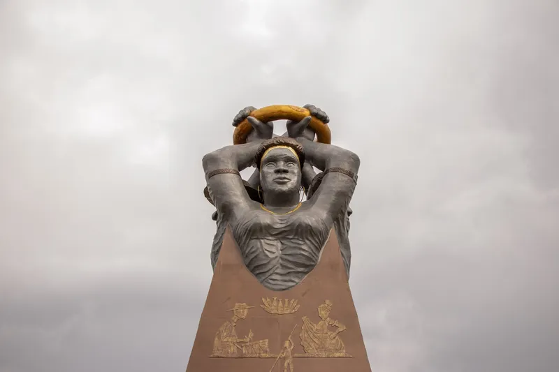 Close-up of the top of the Praça Povos do Meio do Mundo monument: figure with raised arms holding a golden ring, on a base with reliefs.