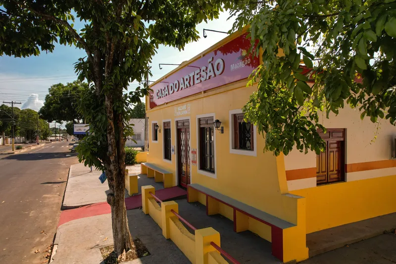 Yellow facade of Casa do Artesão de Mazagão, with a red sign, wooden doors, benches and trees on the sidewalk.