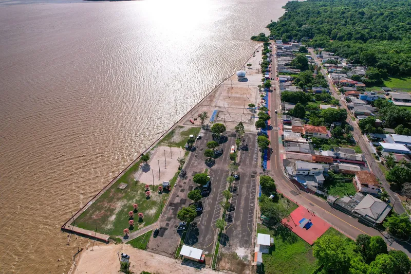 Vista aérea del paseo de la Praia da Fazendinha: estacionamiento arbolado, plaza y calle junto al río Amazonas, con el sol reflejado en el agua.