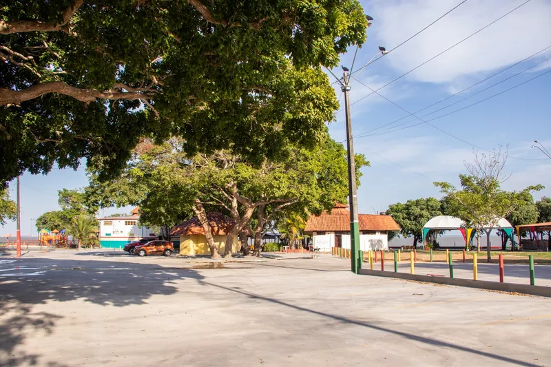 Praça aberta do Complexo do Araxá com grandes árvores, quiosques de telhado de cerâmica, tenda branca e o rio ao fundo.