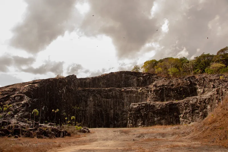 Gran pared de roca oscura de la Montanha da Pluma vista de frente, con árboles en la cima, suelo de tierra y cielo cargado de nubes