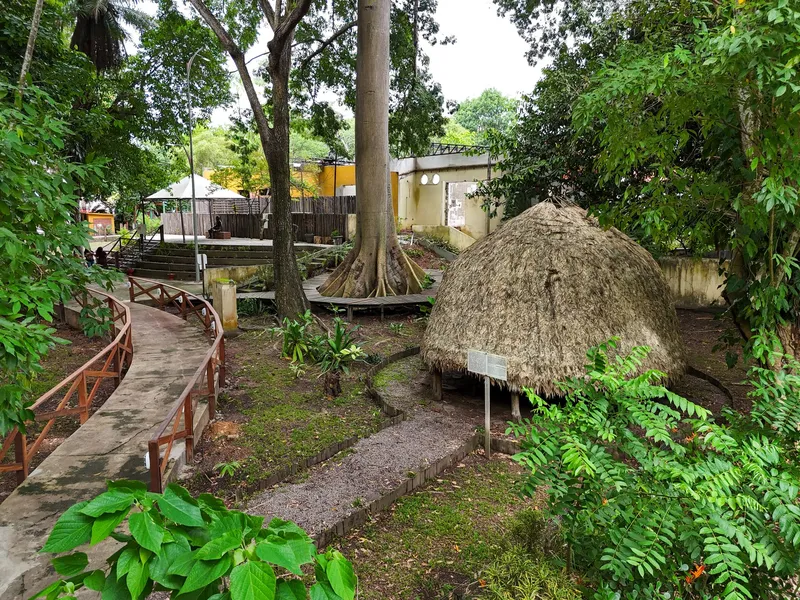 Área exterior arbolada del Museu Sacaca con un camino de concreto, barandas de madera y una casa de paja en forma de cúpula entre la vegetación.