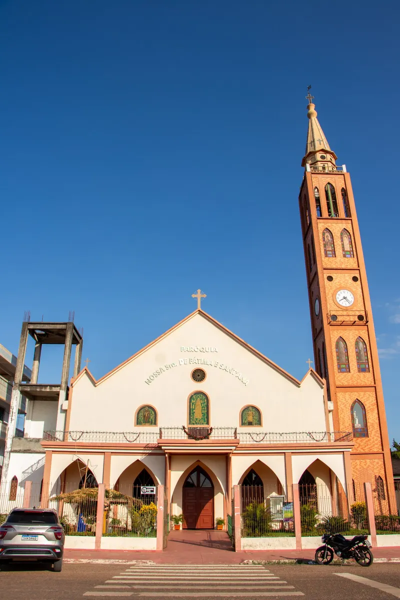 Fachada da Igreja Nossa Senhora de Fátima e Sant'Ana com arcos ogivais e a torre de tijolos com relógio, sob céu azul.