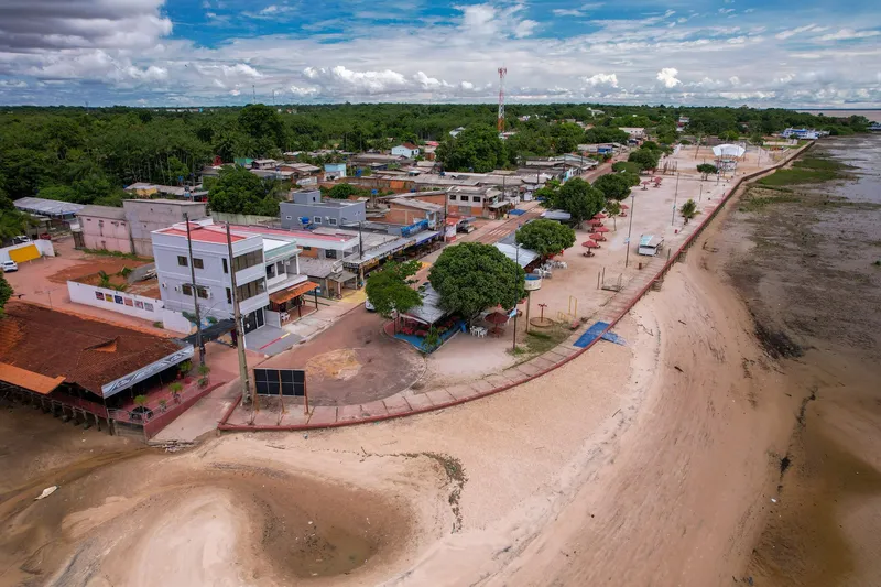 Vista aérea del paseo de la Praia da Fazendinha con marea baja, arena expuesta, murete, quioscos y casas bajo cielo azul con nubes.