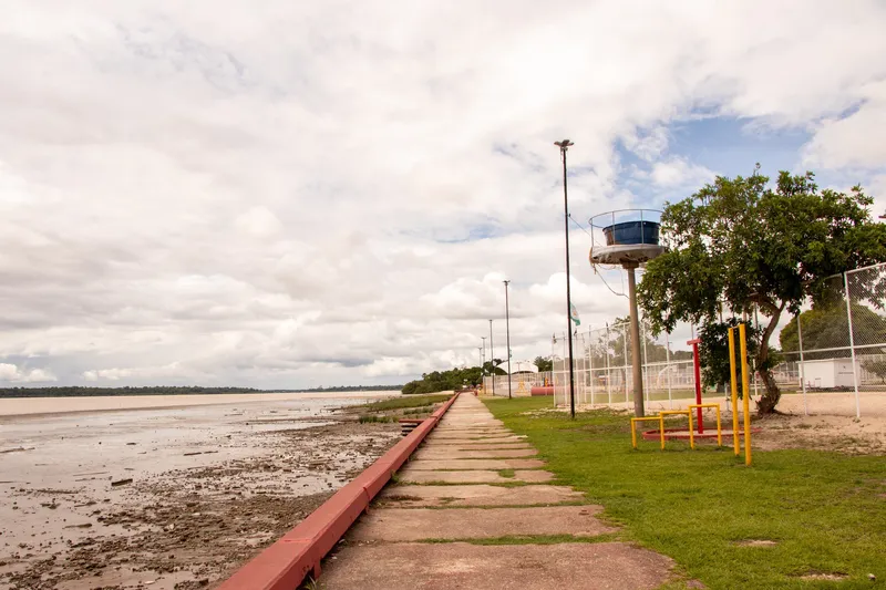 Paseo a orillas del río en la Praia da Fazendinha con marea baja, césped, aparatos de gimnasia y un árbol, bajo cielo nublado.