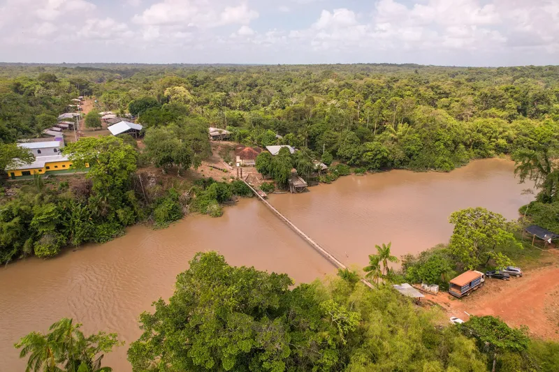 Vista aérea da Vila do Cunani: ponte pênsil sobre o rio barrento ligando as margens, casas entre as árvores e floresta até o horizonte