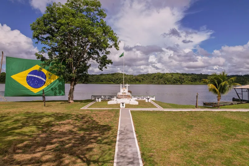 Césped a orillas del río Oiapoque en Clevelândia do Norte, con gran bandera de Brasil, monumento con cruz, mástil y selva en la otra orilla.