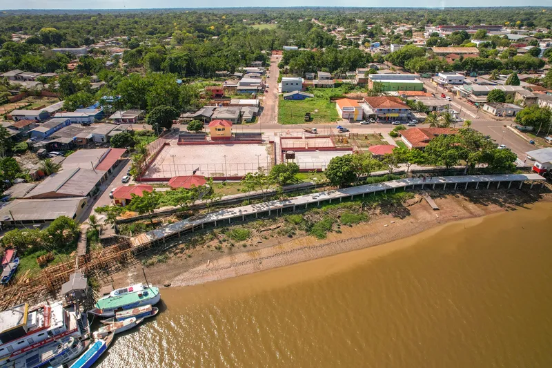 Vista aérea del paseo ribereño de Mazagão Novo: pasarela junto al río, canchas deportivas, barcos amarrados y la ciudad al fondo.