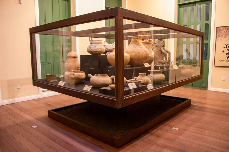 Vitrina de vidrio y madera con vasijas y urnas de cerámica arqueológica en el centro de una sala con piso de madera del Museu Joaquim Caetano da Silva.