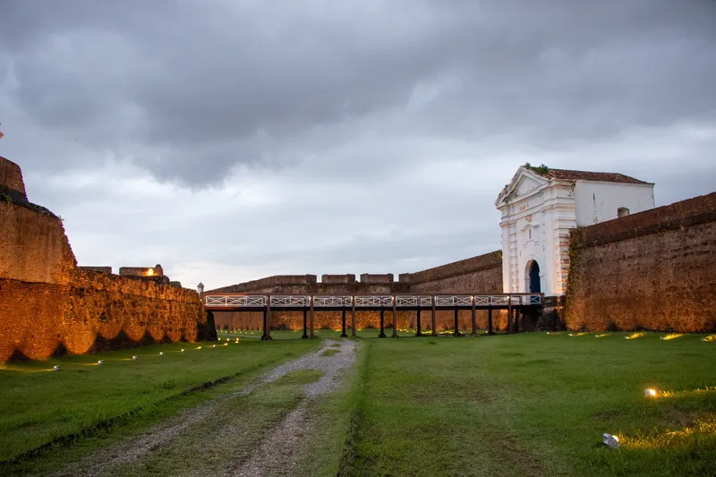 Fosso gramado da Fortaleza de São José de Macapá ao entardecer, com muralhas iluminadas, ponte de madeira e portal branco.