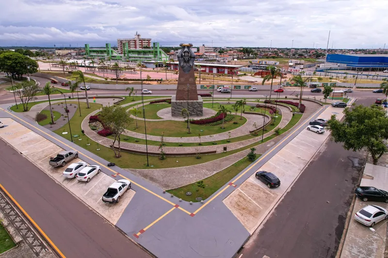 Aerial view of Praça Povos do Meio do Mundo, with the monument, flower beds and a parking area in the foreground, and the city of Macapá in the background.