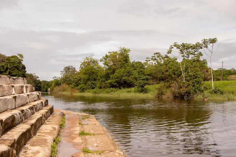 Steps of the Tartarugalzinho waterfront leading down to the calm river, with a bank of trees and grass under a cloudy sky.