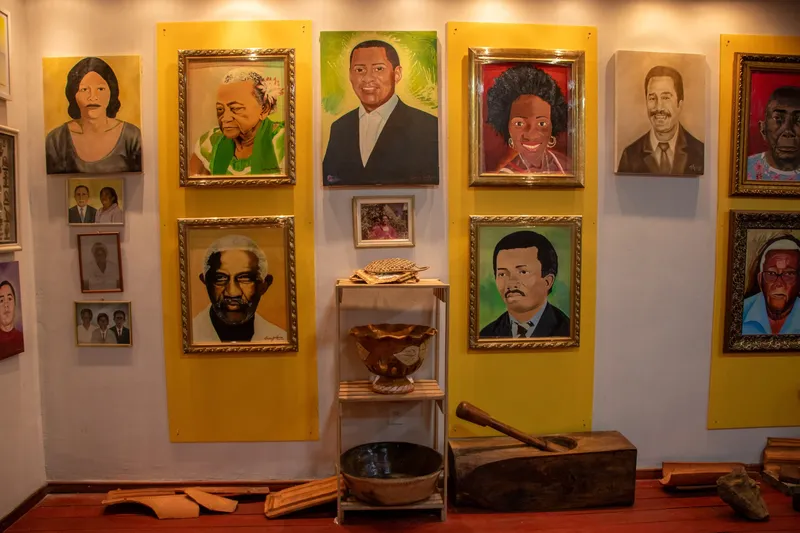Parede com retratos pintados em molduras douradas sobre painéis amarelos no Museu Afro-Amazônico, com objetos de madeira e cerâmica no chão.