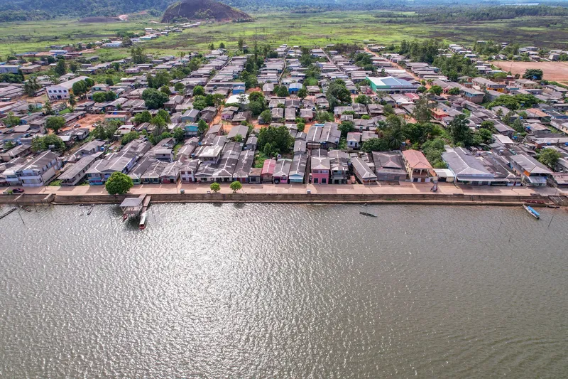 Aerial view of the Vitória do Jari waterfront: a retaining wall along the river, rows of town houses and fields with a hill behind.