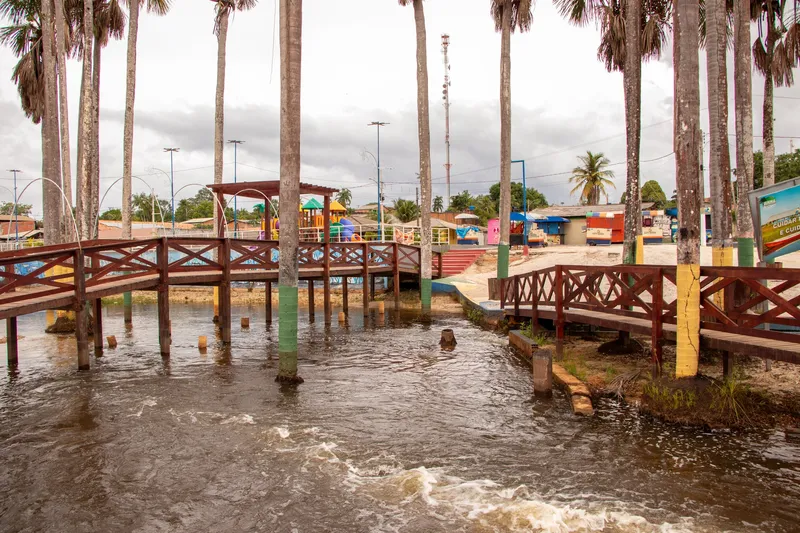 Pasarelas de madera sobre el agua entre palmeras de troncos pintados en el balneario de Porto Grande, con parque infantil al fondo