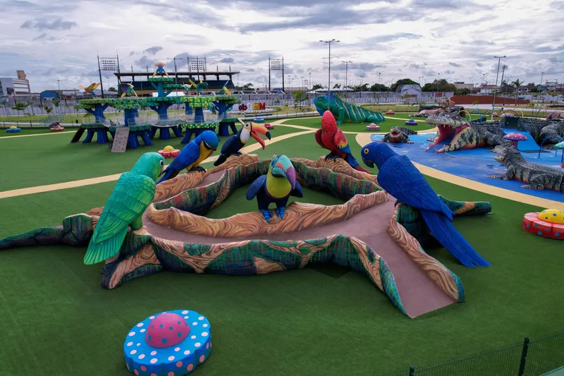 Tobogán en forma de tronco rodeado de esculturas de guacamayos, tucanes y loros coloridos sobre césped sintético en el Parque Meio do Mundo.