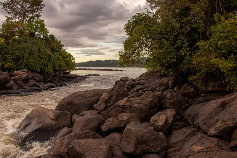 Água agitada da Corredeira Grand Roche passando entre grandes pedras escuras, emoldurada por árvores, com o rio largo ao fundo