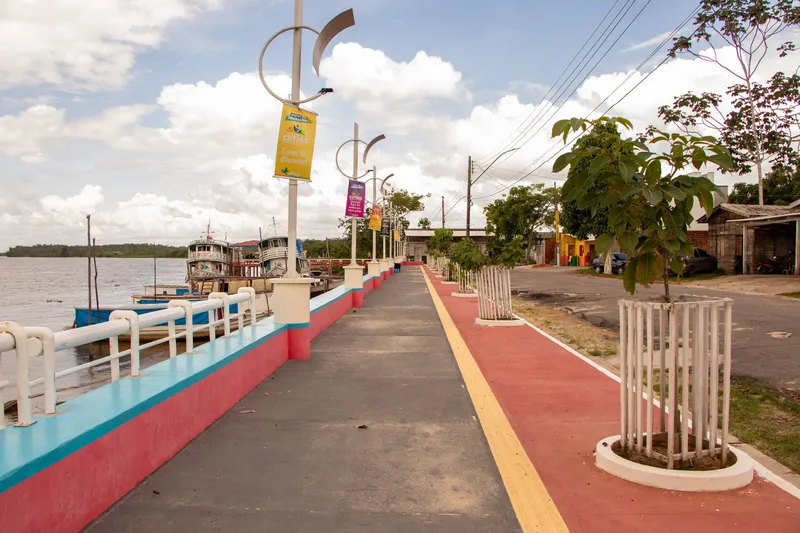 Promenade du front de rivière de Cutias do Araguari avec garde-corps blanc, sol rouge et bleu, lampadaires à bannières et bateaux amarrés