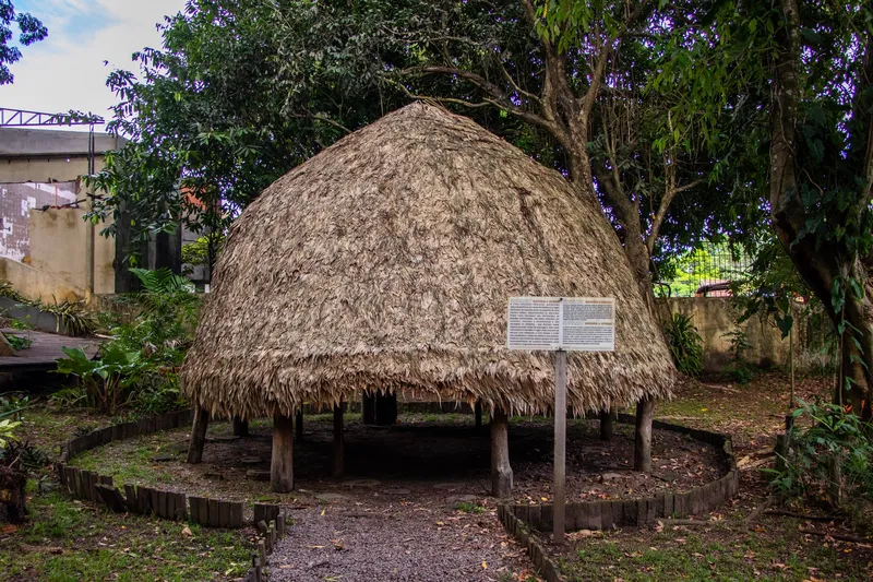 Réplica de casa indígena con techo de paja en forma de cúpula sobre postes de madera y un cartel informativo, en el Museu Sacaca.
