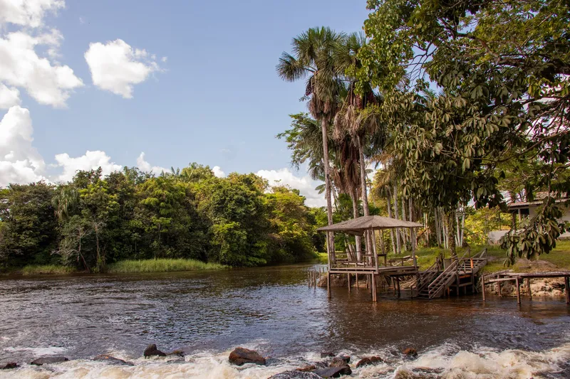 Quiosco de madera con escalera sobre el río en el Balneário Água Fria, palmeras altas y agua corriendo sobre piedras en primer plano