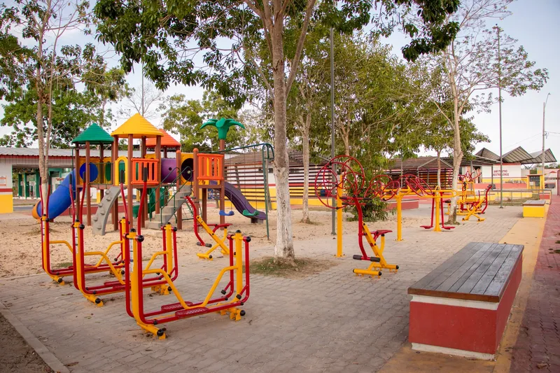 Children's playground and colorful exercise equipment in the Mazagão Novo square, among trees, with a bench in the foreground.