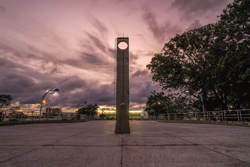 Torre do Marco Zero do Equador ao entardecer, sob céu de nuvens em tons de lilás e rosa, com postes acesos na praça.