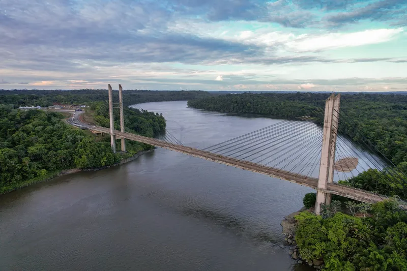 Aerial view of the whole Binational Bridge spanning the Oiapoque River between banks of dense forest, under a cloudy sky