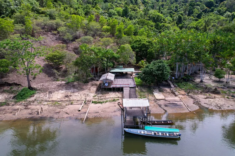 Aerial view of Balneário Recanto do Cupu: a jetty with boats moored on the river, kiosks on the bank and a forest-covered hillside.