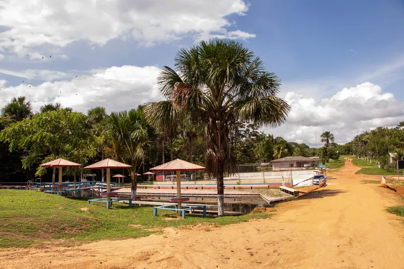 Estrada de terra chegando ao Balneário Sagrado Coração de Maria, com palmeira alta, quiosques de telhado vermelho e deques junto à água