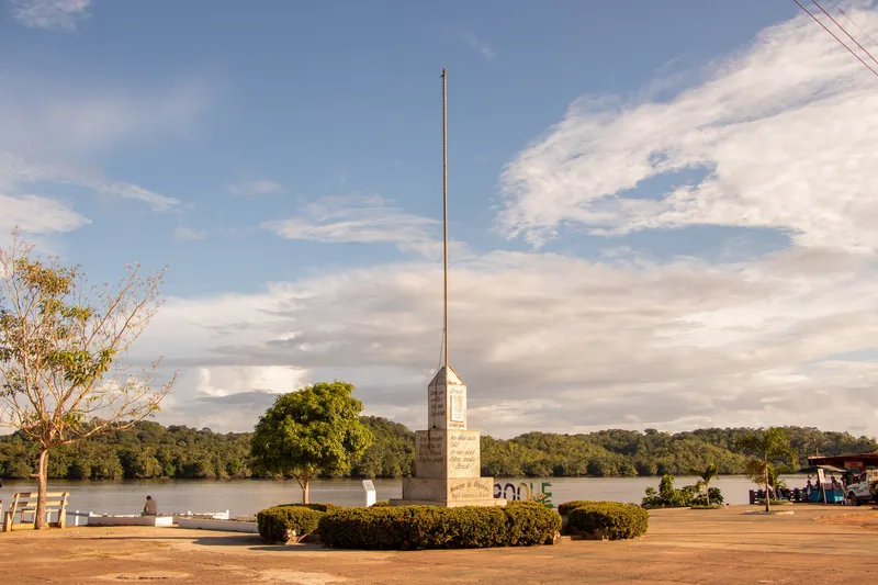 Obelisco del Monumento a la Patria con un mástil alto en la costanera de Oiapoque, rodeado de arbustos, con el río y la selva al fondo