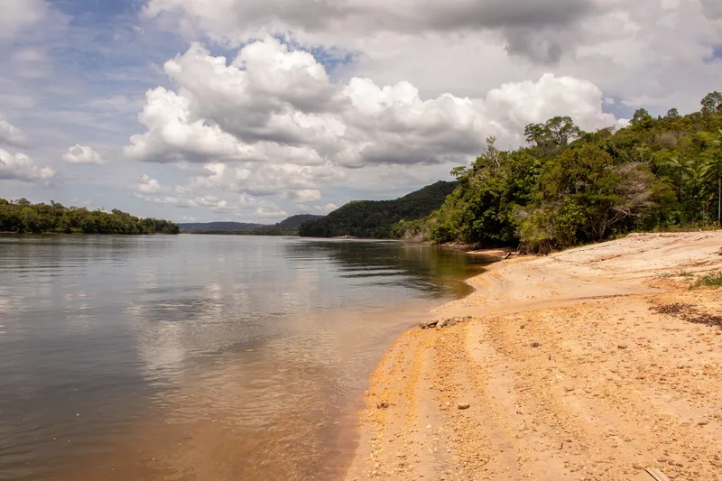 Light sand river beach at Balneário Belas Águas, with forest on the bank, hills in the distance and clouds reflected in the water.