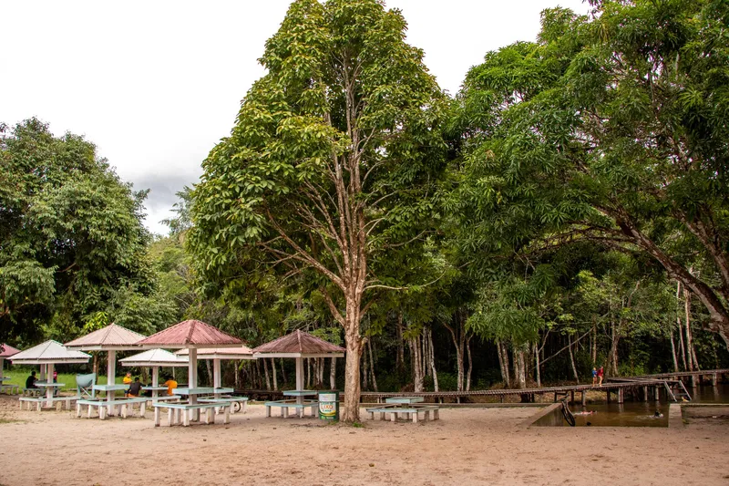 Tables with sunshades on sand under tall trees at Balneário São Joaquim do Pacuí, with a boardwalk over the creek behind.