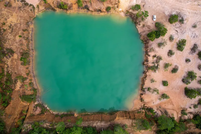 Top-down aerial view of an almost square green lake at Barranco do Geraldão, ringed by pale earth, shrubs and a parked car